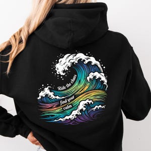 Könnte beinhalten: Schwarzer Kapuzenpullover mit einem farbenfrohen Wellenmuster und dem Text "Ride the chaos find your calm". Das Wellendesign verwendet Blau-, Grün-, Gelb-, Orange- und Lilatöne. Der Hoodie hat eine Kapuze und eine Vordertasche.