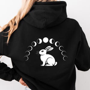 May include: Black hoodie featuring a white graphic of a rabbit and a series of moon phases. The rabbit is centered below the arched moon phase design. The hoodie has a hood and a drawstring.