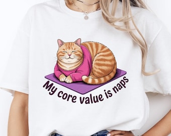 May include: White t-shirt featuring a cartoon cat wearing a pink sweater, lying on a purple mat. The text "My core value is naps" is printed in black and white around the cat.