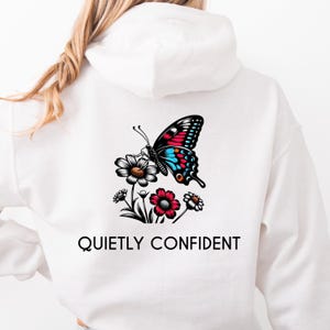 May include: White hoodie featuring a colorful butterfly design with red, blue, and black accents, perched on a cluster of flowers. The text "QUIETLY CONFIDENT" is printed below the graphic.