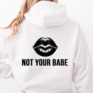 May include: White hoodie with a black graphic of lips and the text "NOT YOUR BABE" in a bold, sans-serif font. The hoodie is made of a soft material and has a hood.