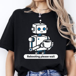 Op de afbeelding: Zwart T-shirt met een wit en blauw robot-ontwerp dat een koffiekopje vasthoudt. De robot heeft een vierkante kop en blauwe ogen. De tekst "Rebooting please wait" staat in een witte rechthoek eronder.
