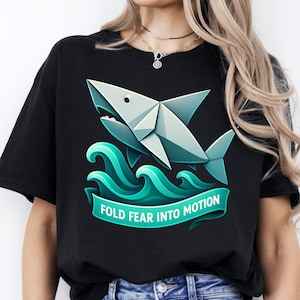 May include: Black t-shirt featuring a geometric origami shark design in shades of gray and teal, emerging from teal waves. A teal banner below reads "FOLD FEAR INTO MOTION".