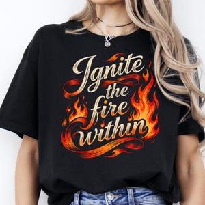 Könnte beinhalten: Schwarzes T-Shirt mit dem Text "Ignite the fire within" in einer stilisierten Schriftart, umgeben von Flammen in Orange- und Rottönen. Ein lässiges Kleidungsstück.