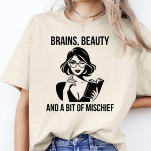 May include: Cream-coloured t-shirt with the text "BRAINS, BEAUTY AND A BIT OF MISCHIEF." The graphic shows a black and white illustration of a woman with glasses holding a book. The woman is wearing a black top.