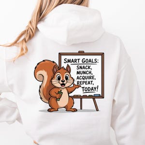 Könnte beinhalten: Weißer Kapuzenpullover mit einem Cartoon-Eichhörnchen und einer Tafel. Auf der Tafel steht "SMART GOALS: SNACK, MUNCH, ACQUIRE, REPEAT, TODAY!". Das Eichhörnchen ist braun und hält einen grünen Gegenstand.