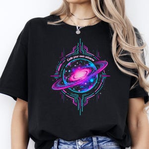 Op de afbeelding: Zwart T-shirt met een levendige afbeelding van een sterrenstelsel in roze, blauwe en paarse tinten, omcirkeld door een ring en geometrische lijnen. De tekst "Code your own universe" staat boven het ontwerp.