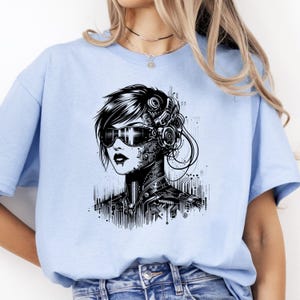 May include: A light blue t-shirt featuring a black and white graphic of a cyborg woman. The design includes a detailed depiction of cybernetic enhancements, including goggles and circuitry, creating a futuristic aesthetic.
