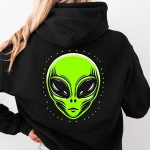 Könnte beinhalten: Schwarzer Kapuzenpullover mit einem neongrünen Alien-Gesicht auf der Rückseite. Der Alien hat große ovale Augen und einen kleinen Mund. Das Design ist von einem gepunkteten Kreis umgeben.