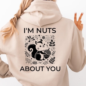Könnte beinhalten: Beigefarbener Kapuzenpullover mit einem schwarz-weißen Grafikdesign eines Eichhörnchens, das eine Eichel hält, umgeben von floralen Elementen. Der Text "I'M NUTS ABOUT YOU" ist oberhalb und unterhalb der Grafik aufgedruckt. Der Hoodie hat eine Kapuze mit Kordelzug.