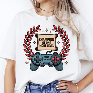 May include: White t-shirt featuring a gaming-themed graphic. The design includes a gamepad, a scroll with the text "CHAMPION OF ONE MORE LEVEL," and a laurel wreath. Accents include gold stars and red leaves. A casual, comfortable tee.