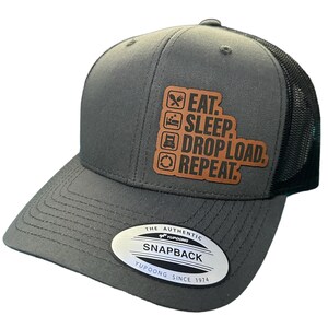 May include: A grey and black trucker hat with a brown leather patch that says "EAT. SLEEP. DROP LOAD. REPEAT." The hat has a black mesh back and a YUPOONG logo on the side.