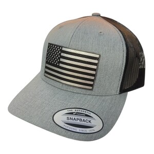 Puede incluir: Una gorra de camionero de malla gris y negra con un parche de la bandera estadounidense en blanco y negro en la parte delantera. La gorra tiene una parte trasera de malla negra y una parte delantera gris con una etiqueta negra "THE AUTHENTIC YUPOONG SNAPBACK YUPOONG SINCE 1974".