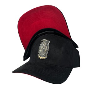 Puede incluir: Gorra de béisbol negra con visera y parte inferior rojas. La gorra presenta un emblema dorado y negro de la Parca. La gorra está hecha de un material suave similar al ante.