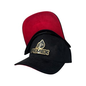 May include: Black baseball cap with a red underside. The cap features a gold-colored emblem with the word "PEMEX". The cap is made of a soft, textured material.
