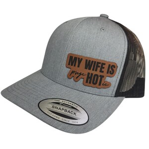 Puede incluir: Gorra de camionero gris y negro con un parche de cuero marrón que dice "Mi esposa es bastante caliente". La gorra tiene una parte trasera de malla negra y una parte delantera gris con una etiqueta de cierre a presión plateada que dice "The Authentic Yupoong Snapback Yupoong Since 1974".