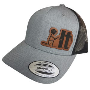 Può includere: Un cappello da camionista grigio e nero con una toppa in pelle marrone che presenta un omino che salda. La toppa dice "I <3 it". Il cappello ha una parte posteriore in rete nera e un'etichetta argentata che dice "The Authentic Yupoong Snapback Yupoong Since 1974".