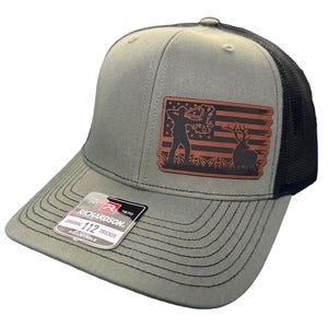 Pode incluir: Um boné de caminhoneiro cinza e preto com um remendo de couro marrom que apresenta a silhueta de uma pessoa com um arco e flecha, um veado e um design de bandeira americana. O boné tem o texto "EST. 1970 RICHARDSON. ORIGINAL 112 TRUCKER ADJUSTABLE" na parte de trás.
