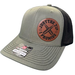 May include: A green and black trucker hat with a brown leather patch that says "Just the Tip I Promise" with a crossed bullet design. The hat is adjustable and has a Richardson 112 Trucker label.
