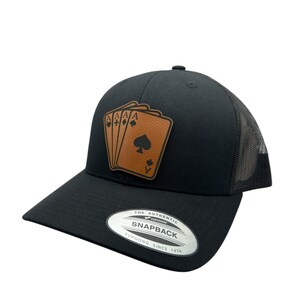 May include: Black trucker hat with a brown leather patch featuring four playing cards, including three aces and a queen of spades. The patch says "A" on each card. The hat has a black mesh back and a silver sticker that says "The Authentic Yupoong Snapback Since 1974".