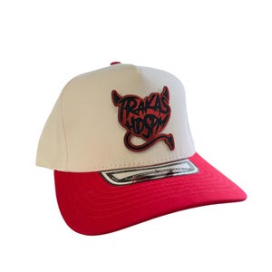 Puede incluir: Gorra de béisbol crema con visera roja. La gorra presenta un gráfico de diablo rojo con el texto "TRAKAS HDSPM". La gorra tiene un ajuste estructurado y una visera curva.