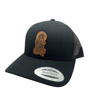 Könnte beinhalten: Schwarze Trucker-Kappe mit einem braunen Leder-Patch, der ein religiöses Bild und den Text "Jesus Saves" zeigt. Die Kappe hat ein Netzrücken und ein YUPOONG SNAPBACK-Etikett.