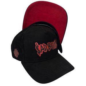 May include: Black and red baseball cap with the text "SAD BOY" in red on the front. The cap has a red underside.