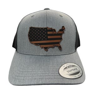 May include: A grey and black trucker hat with a brown leather patch of the United States with a black and white American flag design. The hat has a black mesh back and a white label that says "The Authentic Yupoong Snapback Yupoong Since 1974".