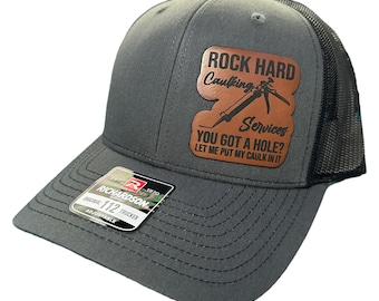 May include: A gray and black trucker hat with a brown leather patch that says "Rock Hard Caulking Services. You got a hole? Let me put my caulk in it."