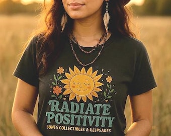 May include: A woman wearing a brown hat, feather earrings, and a dark green t-shirt with a smiling sun graphic and the words "RADIATE POSITIVITY." She is also wearing beaded necklaces and bracelets. The background is a field of tall grass.