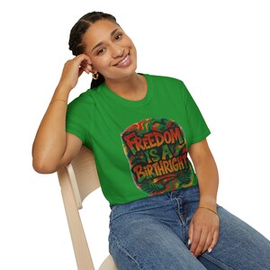 May include: A green t-shirt with the text "FREEDOM IS A BIRTHRIGHT" in red and yellow lettering. The graphic has a colorful, textured design with red, yellow, and green accents. The shirt is paired with blue jeans.