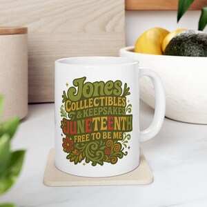 May include: White ceramic mug with the text "Jones Collectibles & Keepsakes Juneteenth Free to Be Me" in green, orange, and yellow. The mug has a handle and is on a beige coaster.
