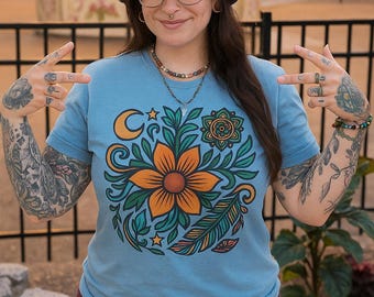 May include: Light blue t-shirt featuring a large floral design with an orange flower, green leaves, and a crescent moon. The shirt is paired with burgundy pants and a patterned bucket hat. The design has a bohemian aesthetic.