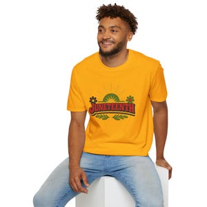 May include: Mustard yellow t-shirt with the word "Juneteenth" in a retro design. The design includes a sunburst, flowers, and leaves. The shirt is worn by a person wearing blue jeans.
