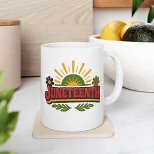 May include: White ceramic mug featuring the word "Juneteenth" in a colorful design. The design includes a green and yellow sun, flowers, and leaves. The mug is sitting on a beige coaster.