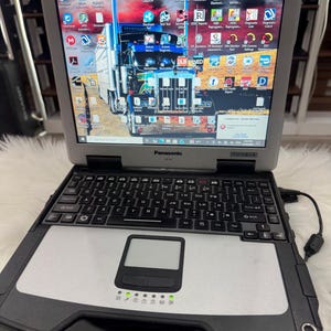 May include: A Panasonic Toughbook CF-31 laptop with a black and silver design. The screen displays a truck image and various software icons. The keyboard and touchpad are visible, along with indicator lights.