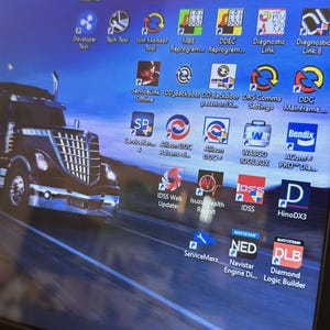 Könnte beinhalten: Der Bildschirm eines Laptops zeigt einen Desktop mit verschiedenen Symbolen, darunter Softwareanwendungen für die Wartung und Reparatur von LKW. Die Symbole sind in einem Raster angeordnet und beinhalten Namen wie "ServiceLink", "Allison DOC", "WABCO TOOLBOX" und "HinoDX3". Das Hintergrundbild ist ein verschwommenes Foto eines schwarzen Sattelzuges, der auf einer Autobahn fährt.