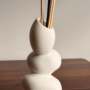 May include: A white ceramic vase with a stacked, pebble-like organic shape. The vase features a textured surface and holds several thin, straight reeds in black and light tan colors, extending upward from the narrow opening at the top.