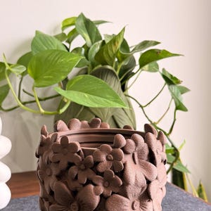 May include: A brown cylindrical planter featuring a textured exterior surface covered in three-dimensional floral shapes. The word TerraBloom is printed in a light pink font above the planter. The planter has a wide opening at the top.