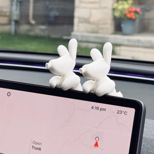 Puede incluir: Dos figuras blancas con acabado mate en forma de conejo están posadas en el borde superior de la pantalla de infoentretenimiento de un vehículo. La pantalla muestra un mapa de navegación con el texto "Open Trunk", "4:18 pm" y "23°C".