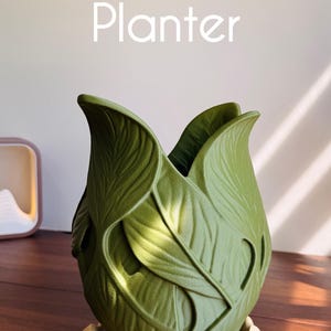 Puede incluir: Una maceta verde LumiLeaf Planter con una superficie texturizada que presenta un diseño de hojas superpuestas. La maceta tiene una abertura ancha con bordes curvos y se asienta sobre una base festoneada clara. El texto LumiLeaf Planter es visible.