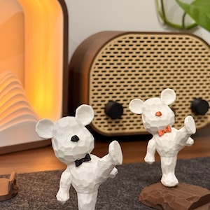 May include: Two white, low-poly bear figurines standing on rectangular bases. The bear in the foreground has a black nose and bowtie, standing on a blue base. The bear in the background has an orange nose and bowtie, standing on a brown base.