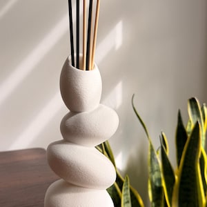 May include: A white reed diffuser with a stacked stone design, featuring the text STILLSTONE Diffuser at the top. The vessel is composed of four rounded, pebble-like shapes stacked vertically, with several thin black and tan reeds inserted inside.