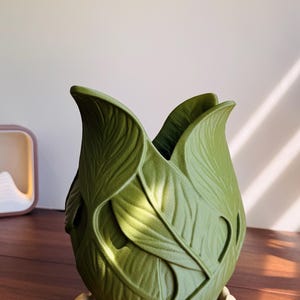 Puede incluir: Un jarrón verde oliva mate con un patrón de hojas superpuestas y texturizadas en su superficie exterior. El jarrón tiene una abertura ancha y festoneada en la parte superior y descansa sobre una base circular separada de color crema claro.
