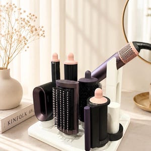 May include: A white hair styling tool organiser stand holding a dark purple and pink hair dryer with multiple attachments including a paddle brush, a round brush, and two curling barrels. The stand is square with dedicated slots for each component.