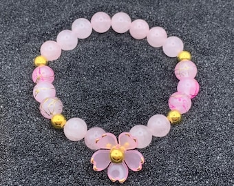 May include: A pink and gold bracelet with a flower charm. The bracelet is made of pink beads with gold accents and a pink flower charm with gold accents.