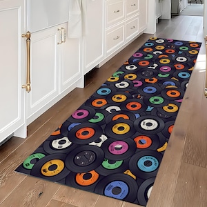 May include: A rectangular rug featuring a pattern of overlapping vinyl records in various colors, including orange, blue, green, and pink. The rug has a dark background and is placed on a wooden floor, suitable for a kitchen or hallway.