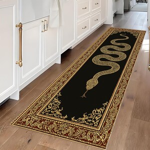 Op de afbeelding: Een rechthoekige zwarte loper met een gouden slangenmotief. De loper heeft een gouden en bordeauxrode rand met sierlijke details. De slang is goudkleurig en de loper ligt op een houten vloer.
