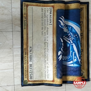Może przedstawiać: Złożony prostokątny dywan z motywem karty Blue Eyes White Dragon. Karta ma niebieską ilustrację smoka, tekst i tekst "Blue Eyes White Dragon". Dywan ma r&oacute;wnież tekst "ATK/3000 DEF/2500".