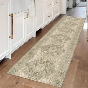 Könnte beinhalten: Ein rechteckiger Teppich mit Muster in einer Küche. Der Teppich hat ein verblasstes Vintage-Design mit einer Farbpalette aus Creme und Salbeigrün. Er liegt auf einem Holzboden, mit weißen Schränken und goldfarbener Hardware im Hintergrund.